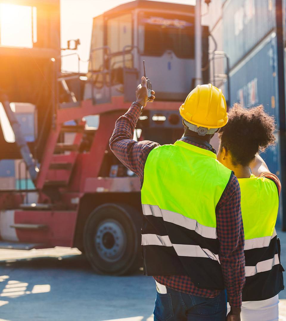 Two workers in hi-vis and hard hats at a container truck in a port yard.