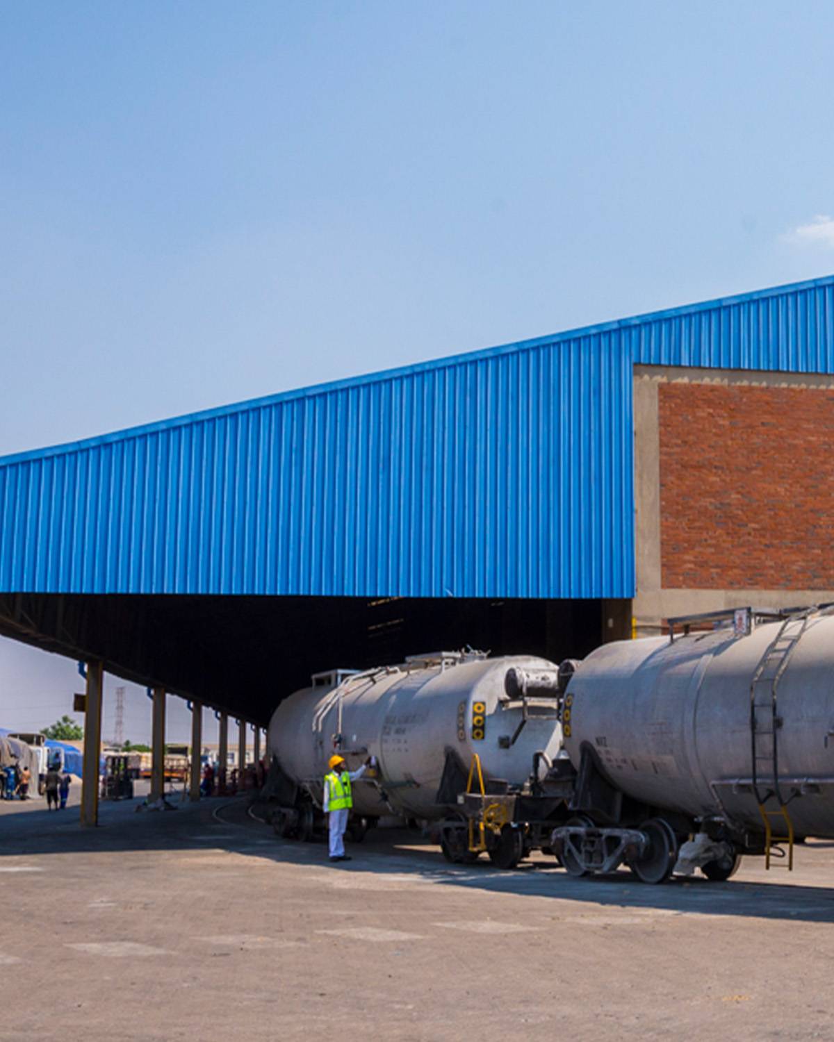 Rail tanker wagons under a blue warehouse canopy, with a worker in hi-vis on the platform.