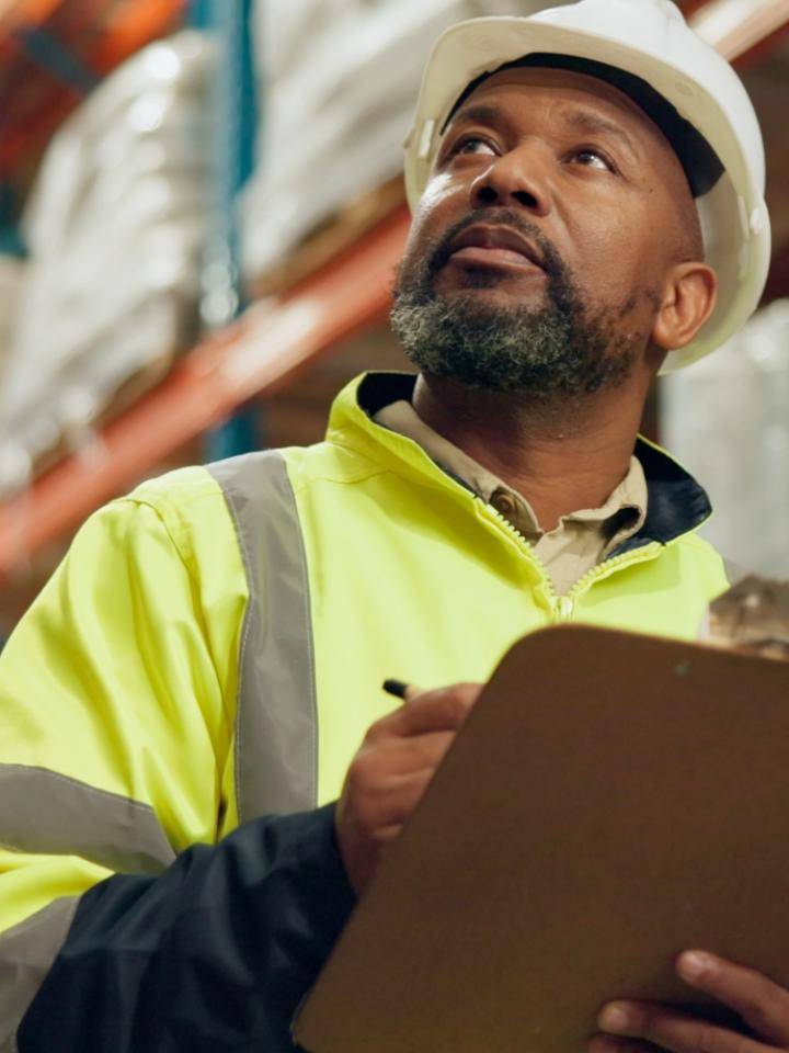A supervisor in a hard hat and hi-vis vest writing on a clipboard in a warehouse.